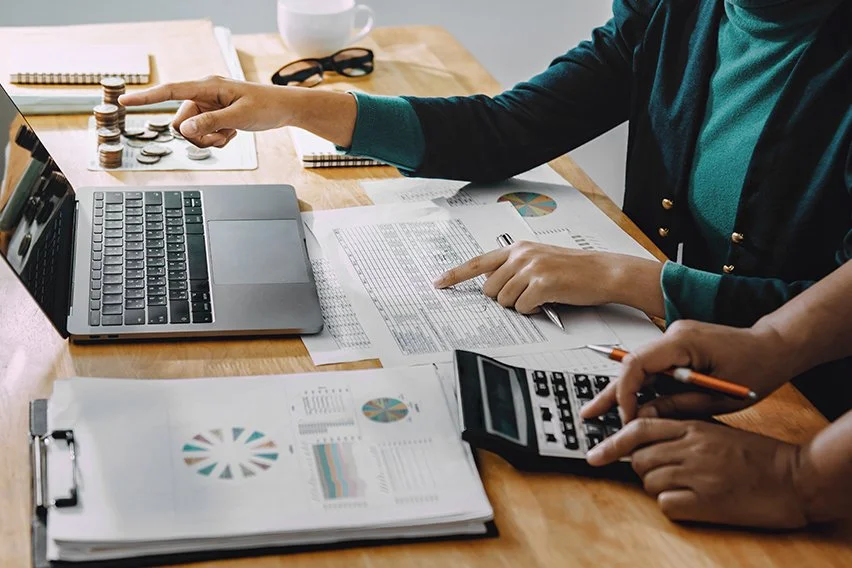 Two people working with documents and a laptop, using a calculator and pointing at charts and spreadsheets on a desk in canada